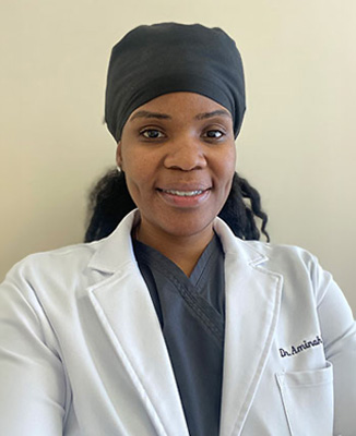 A woman in a white lab coat stands against a wall with a smile, wearing a black headscarf and holding a stethoscope.