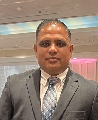 The image features a man standing in an indoor setting, wearing a suit and tie, with a neutral expression.