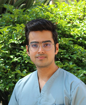 The image shows a man wearing glasses, with a beard and mustache, posing in front of a green hedge. He is dressed in a light-colored shirt and dark pants, and has short hair.