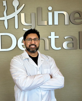 The image shows a man standing in front of a sign that reads  HYLINE DENTAL.  He is wearing a white lab coat and has his arms crossed.