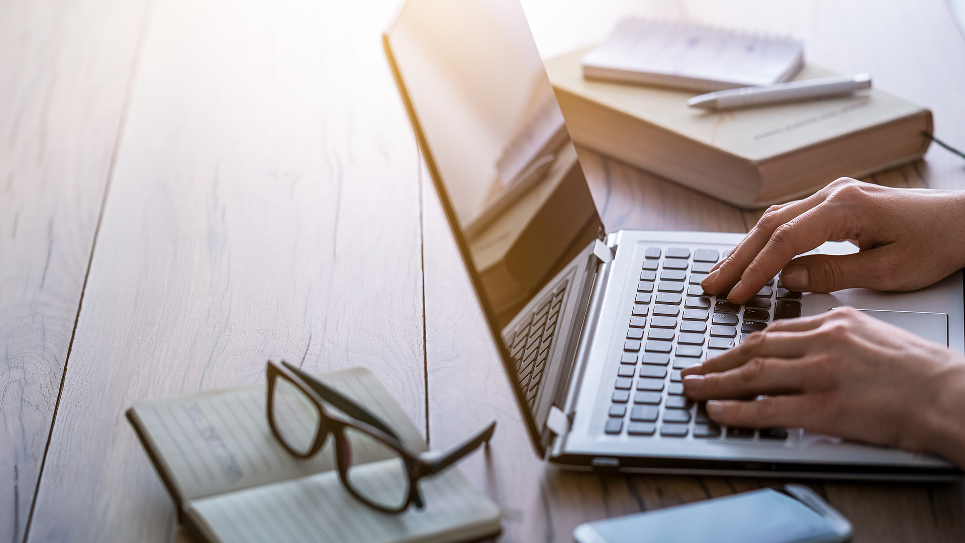 An individual working on a laptop at a desk with books and a pen, set against a blurred background.