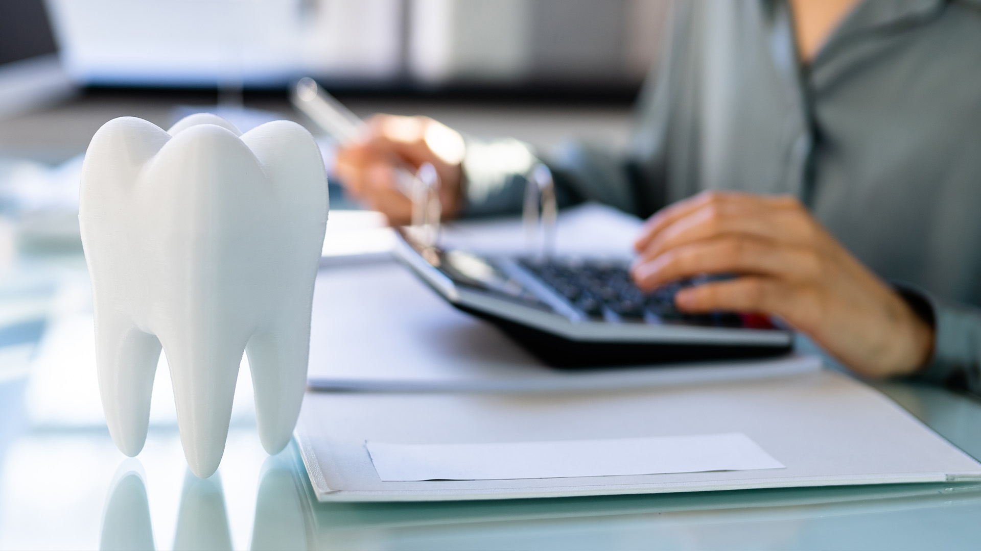 The image shows a person using a laptop at a desk, with a 3D printed model of a tooth and a calculator in the foreground.