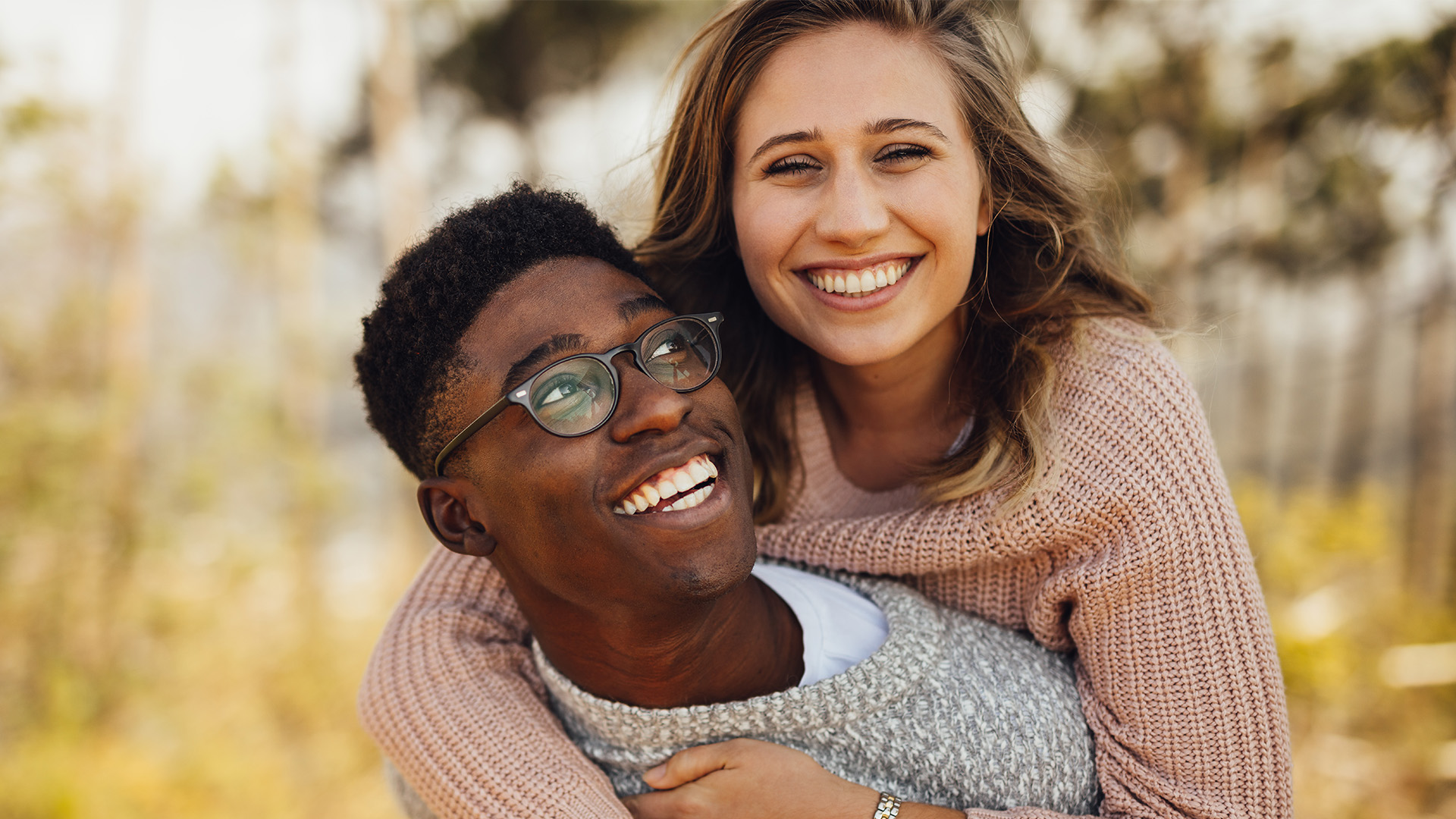 A man and woman embracing, smiling, with one wearing glasses and the other a sweater, posing outdoors.