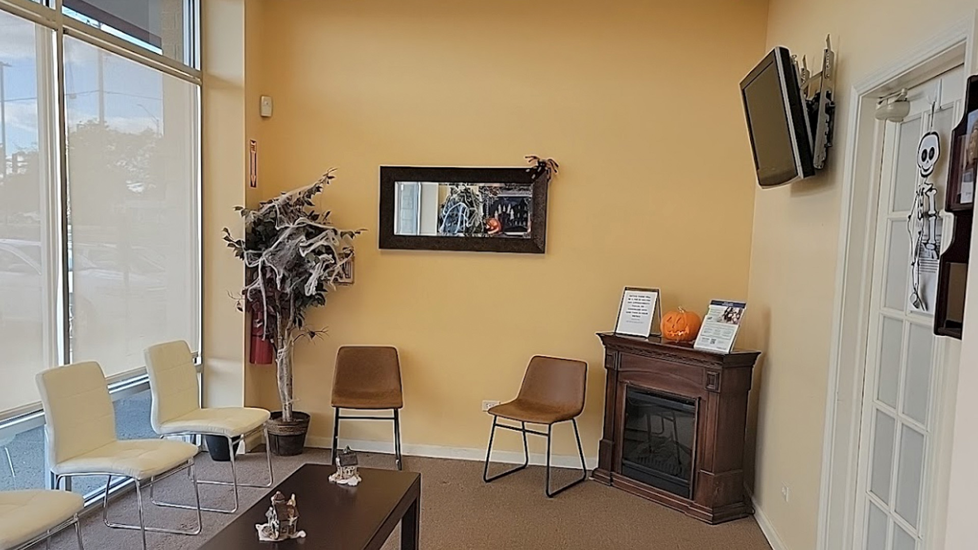 The image depicts an interior space of a room, likely a waiting area or lobby, with a television mounted on the wall displaying static. There is a reception desk in the center with chairs and a small table. A framed picture hangs above the desk. The room has a beige wall and a large window to the left.