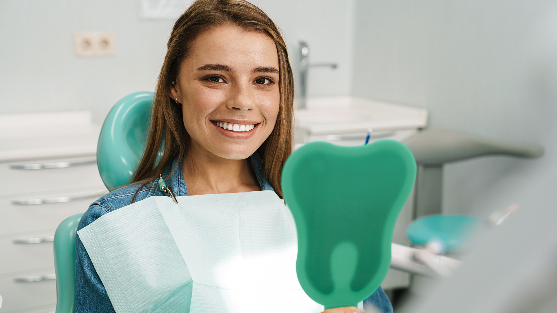 A young woman in a dental office, smiling and holding up a green toothbrush.