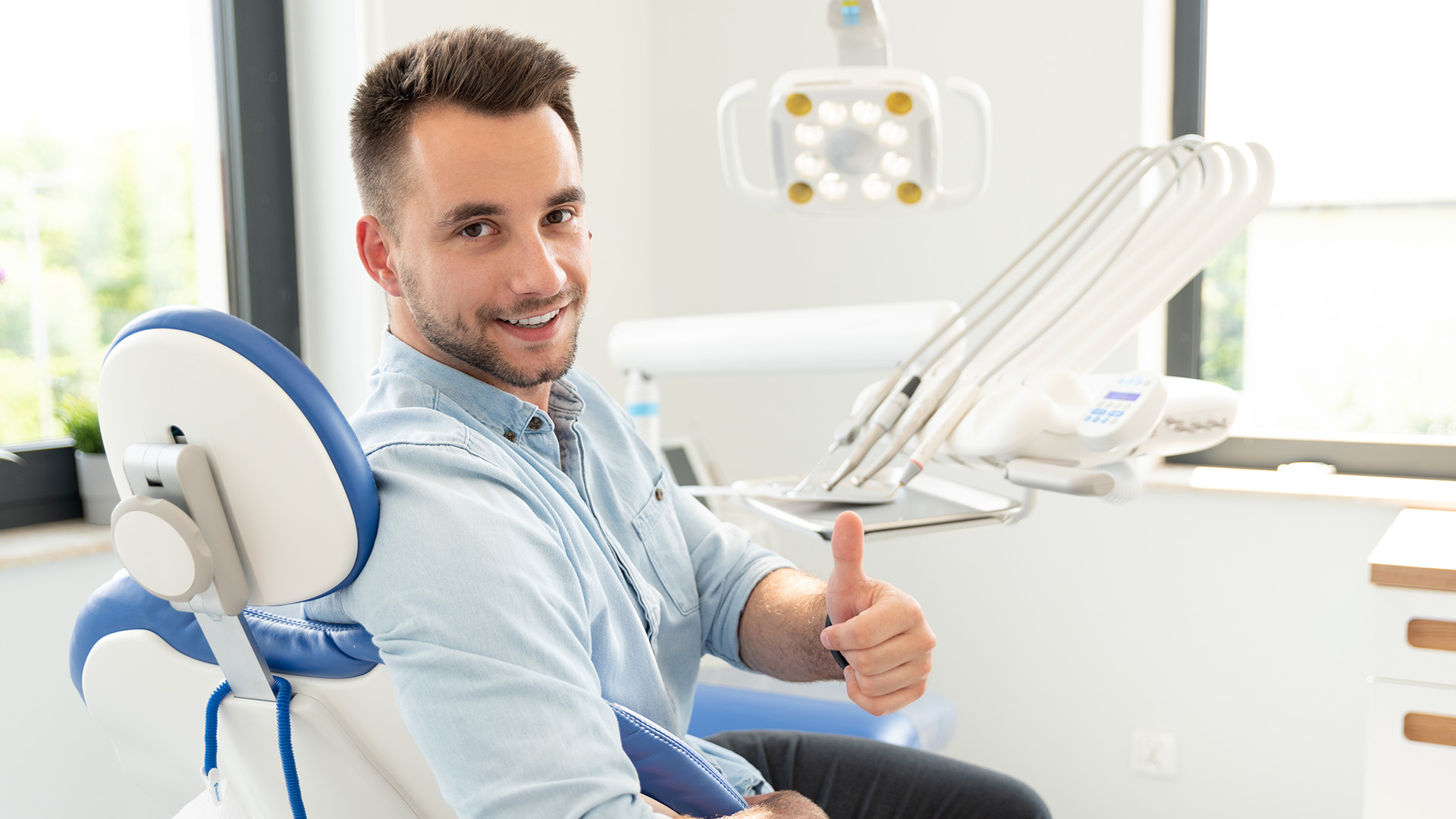 A man in a dental office, smiling and giving thumbs up, sitting in a dental chair with dental equipment around him.