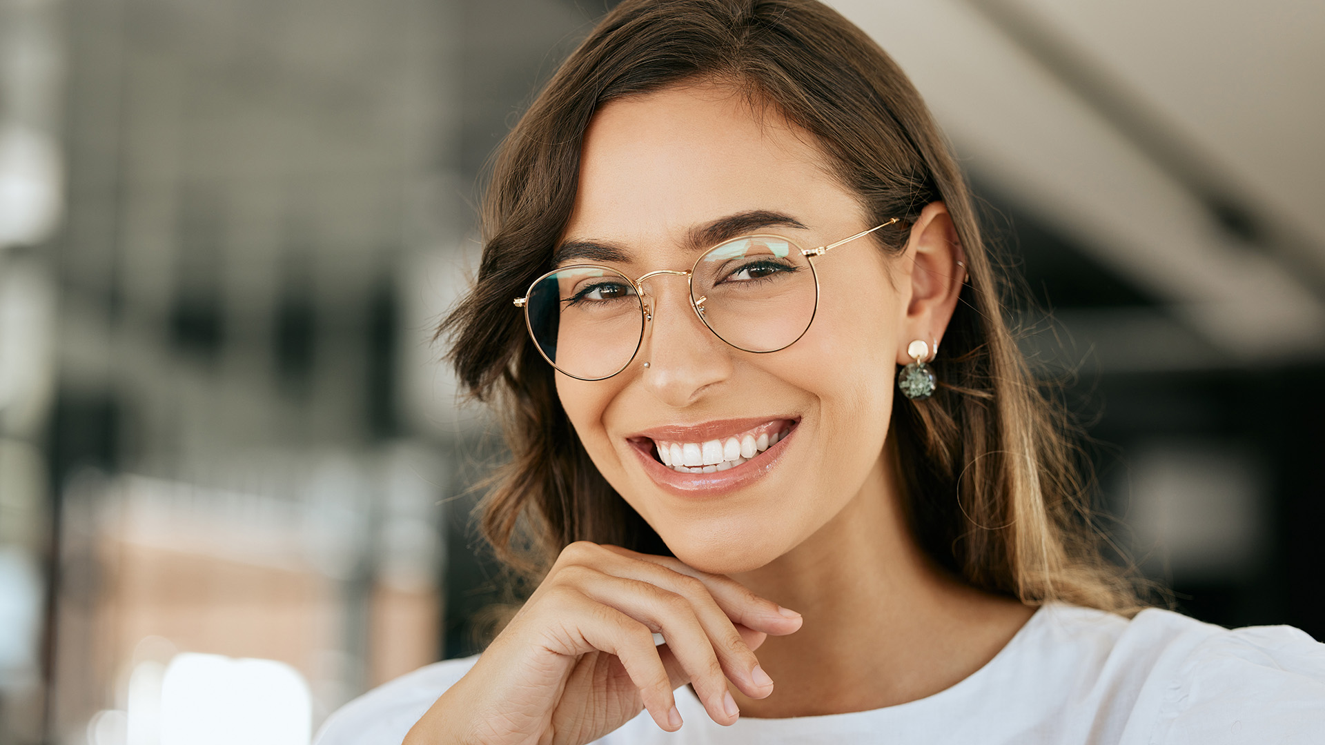 A woman with glasses is smiling at the camera, wearing a white top and sitting in front of a mirror.