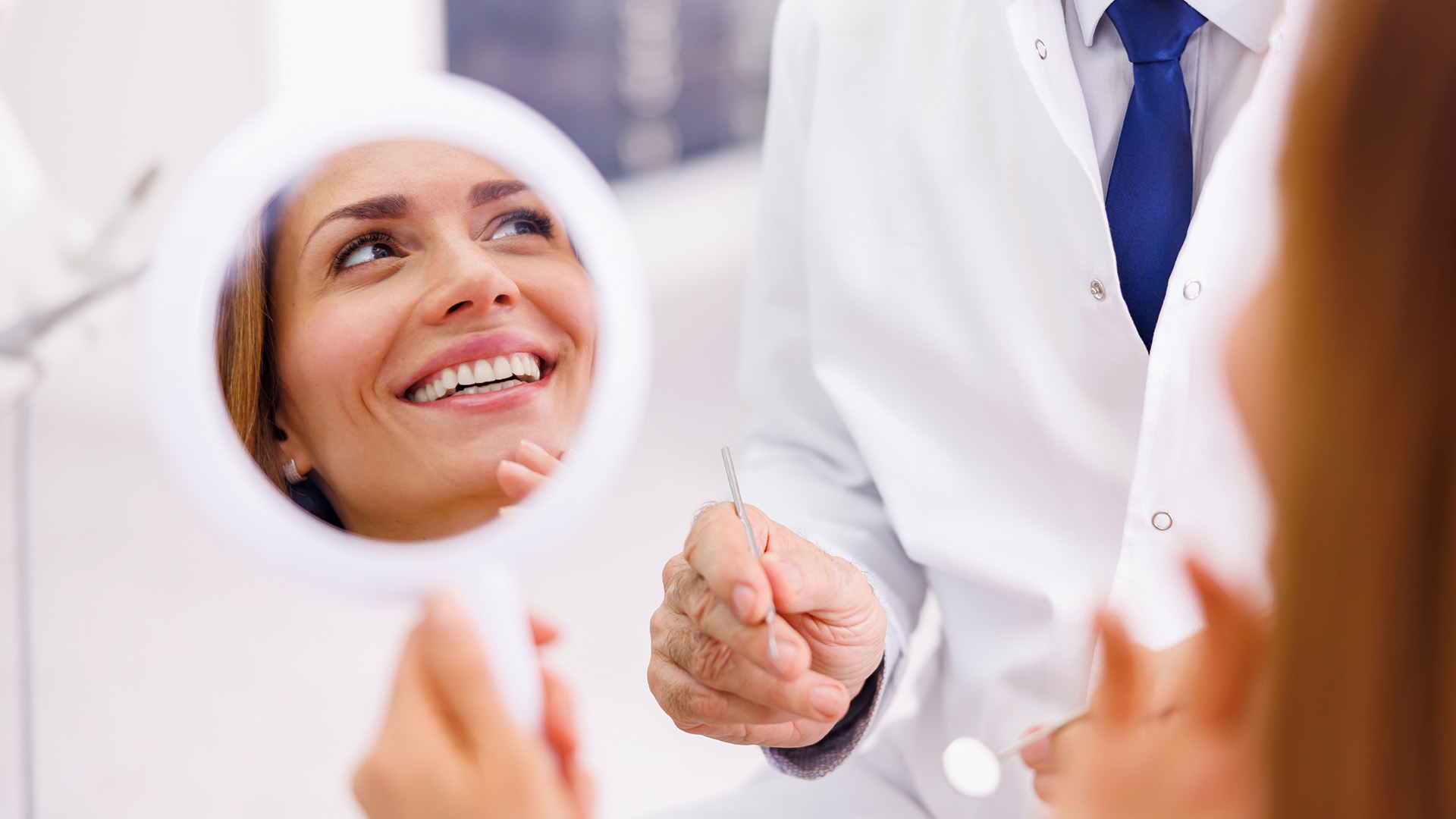 A dentist in a professional setting, smiling at the camera while holding a mirror to show the reflection of a patient s teeth.