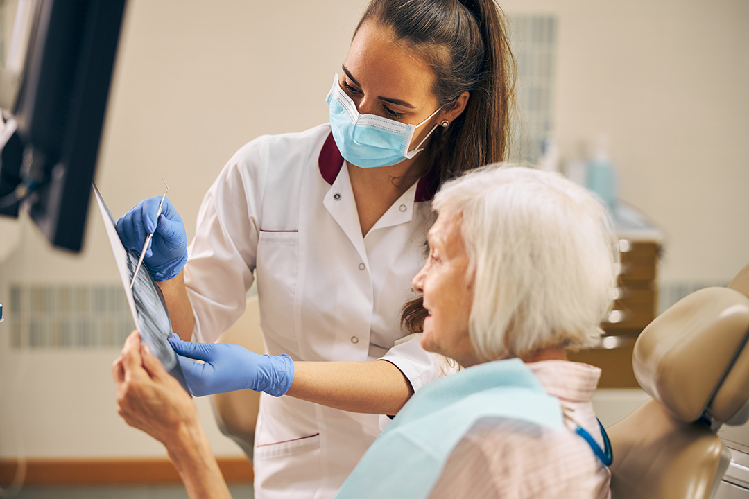 Woman in a white coat is assisting an elderly woman with a dental appointment, both wearing masks and gloves.