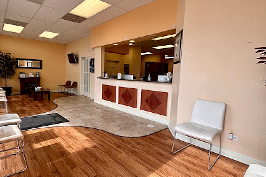 The image depicts an interior view of a spacious room with a wooden floor, featuring a reception area with a counter and chairs, and a waiting area with a couch and chairs.