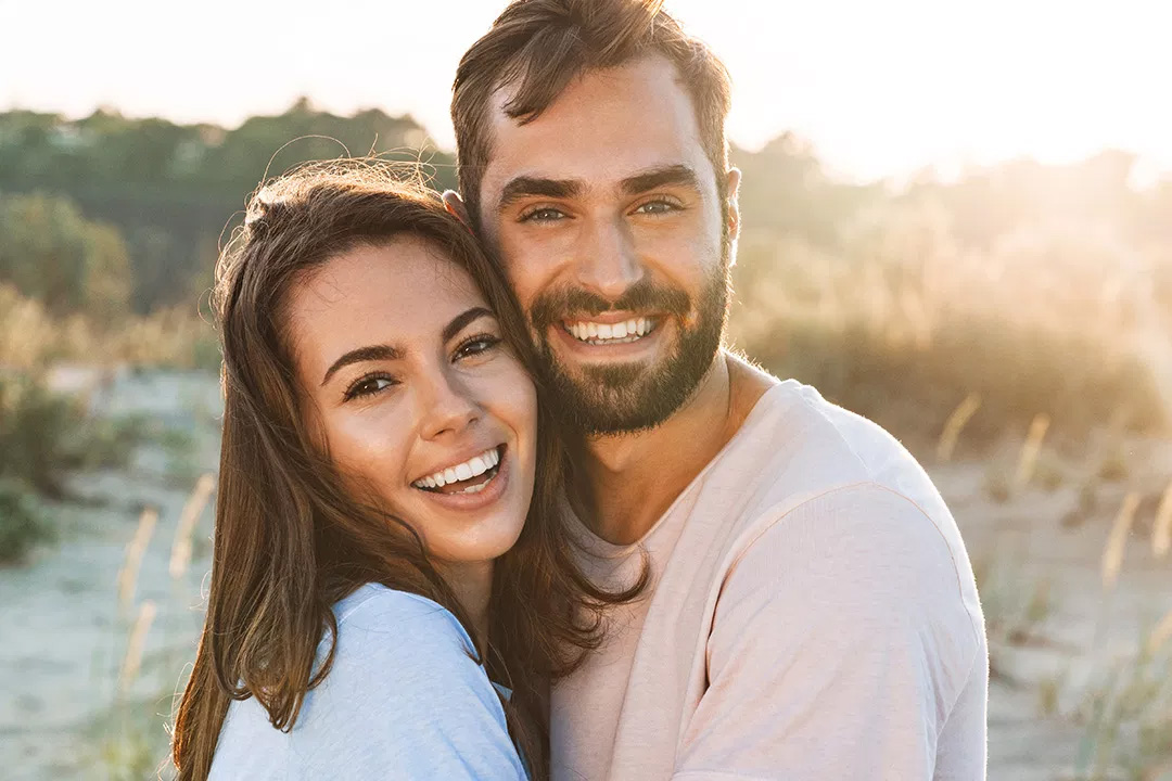 A couple embracing in a sunlit outdoor setting, with the man wearing a beard and both smiling at the camera.