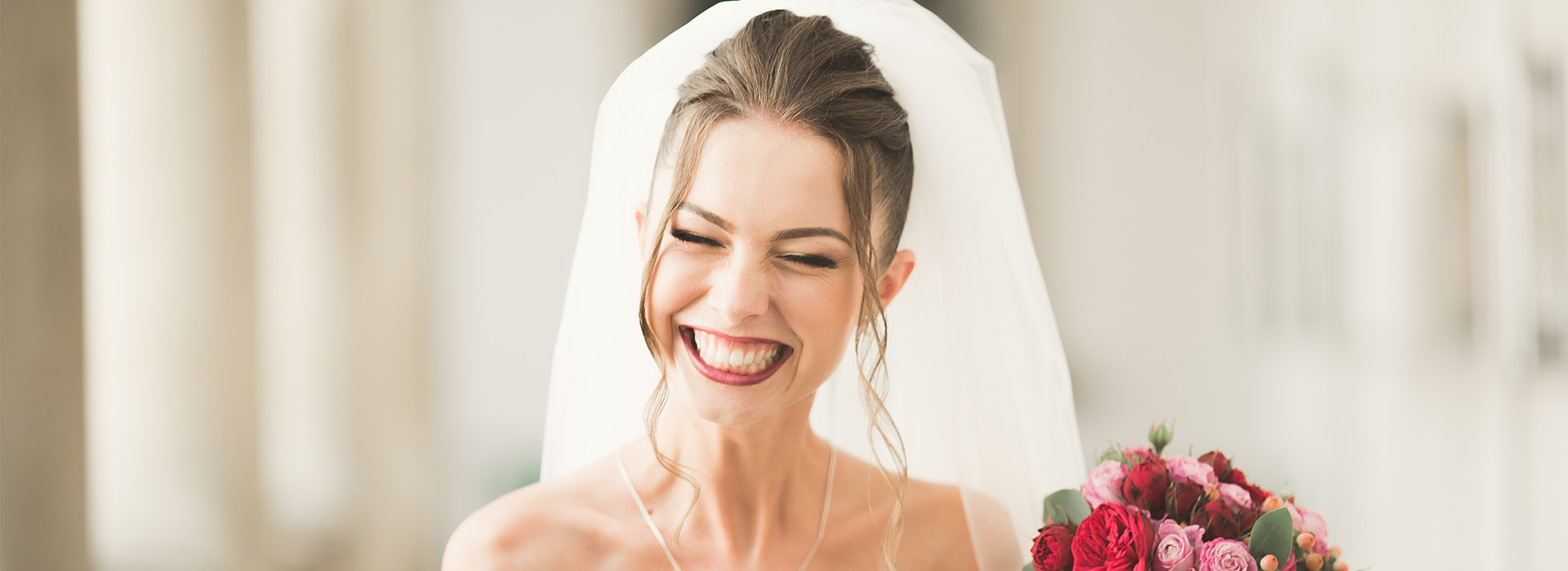 A bride in a white wedding dress, smiling and holding a bouquet, with her groom behind her.