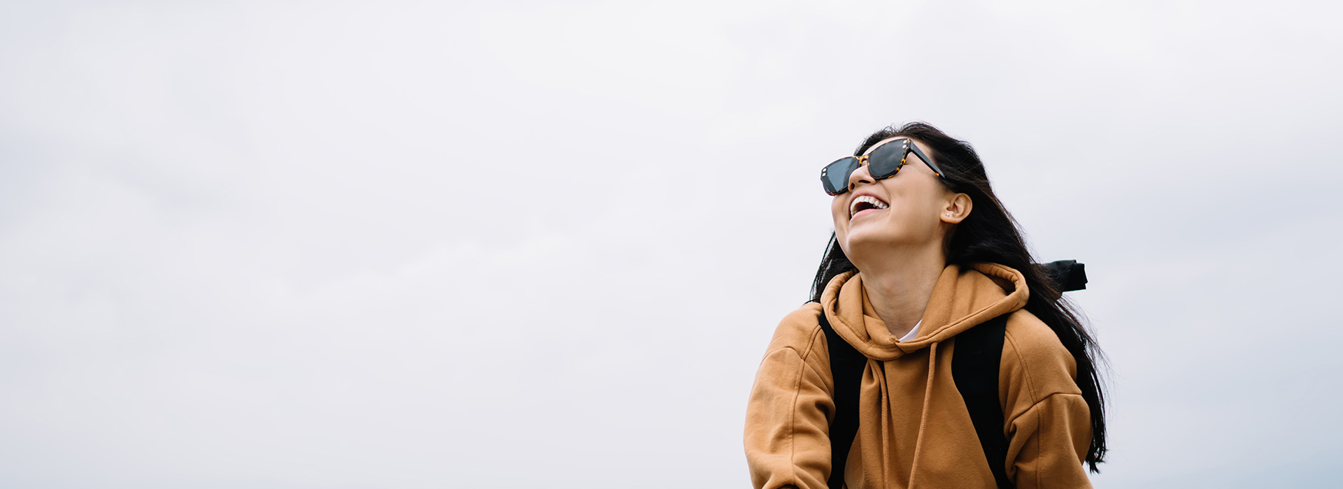 The image is a photograph of an individual wearing sunglasses and a backpack, standing outdoors with a clear sky in the background.