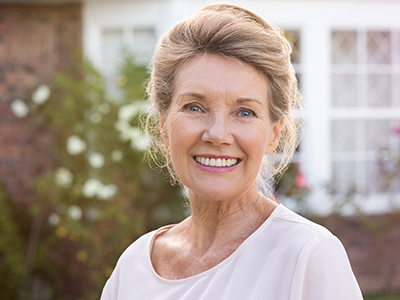 A smiling woman with short hair, wearing a white top and standing in front of a house.