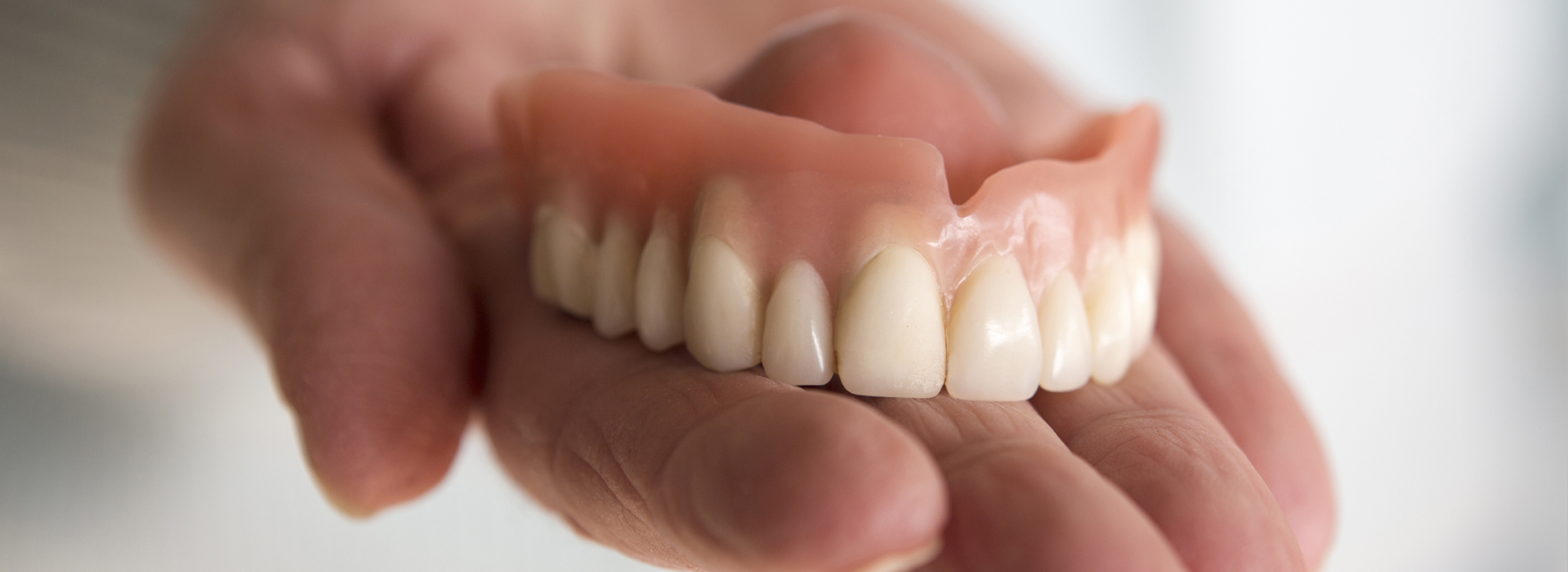 An adult human hand holding a single denture with visible teeth and gumline.