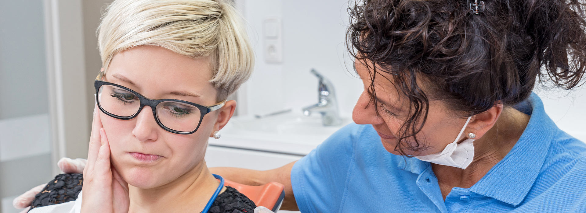 The image shows a woman with glasses and a short haircut receiving dental care, possibly a cleaning or checkup, from another woman who appears to be a dental professional.