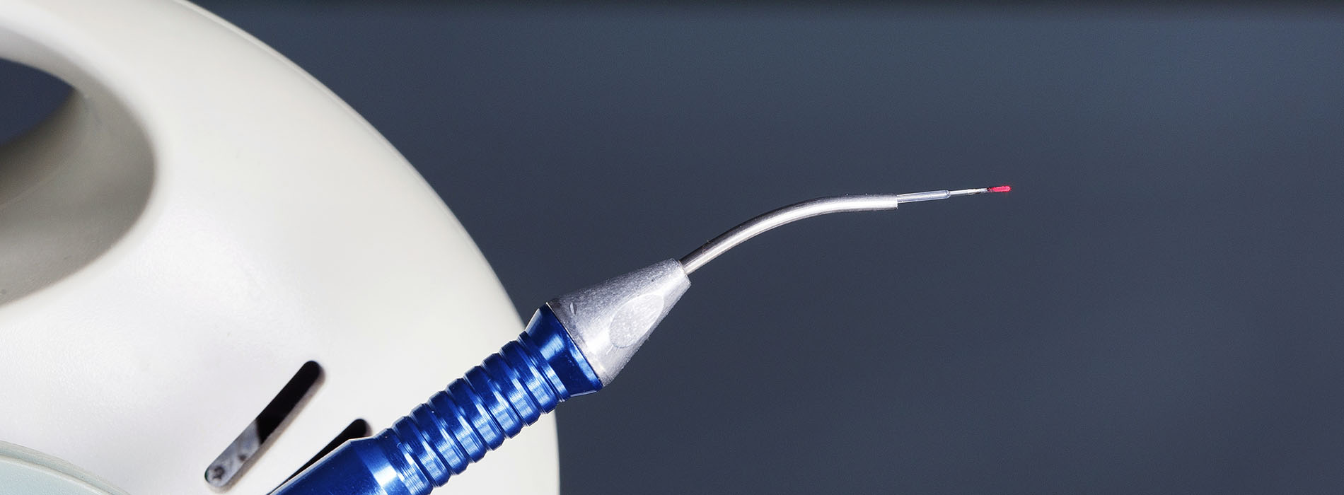 The image displays a close-up of a medical device with a needle and thread, set against a blurred background that includes what appears to be a medical device or instrument.