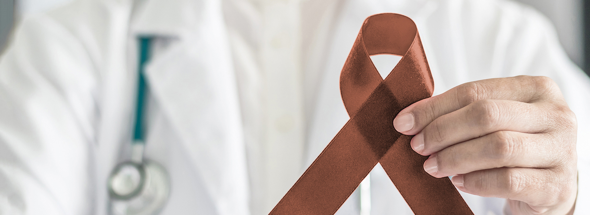 The image shows a person wearing a white coat, holding up a red ribbon with a brown bow on it.