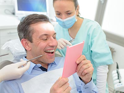 A smiling man in a dental chair, holding up a pink card with a surprised expression, while a woman in scrubs looks on.