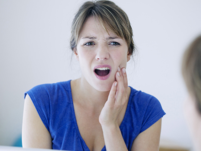 A woman with her mouth open, looking at her reflection in a mirror, appears to be brushing her teeth or washing her face.