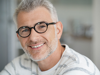 The image shows a man with glasses and a beard, smiling at the camera. He has short hair and is wearing a white shirt with a patterned collar. The background is blurred but appears to be an indoor setting with natural light.