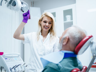 A woman in a white lab coat is assisting an elderly man with dental equipment.