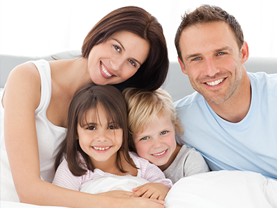 The image shows a family of four, including an adult couple and their two children, posing together in a bedroom with a white wall in the background.