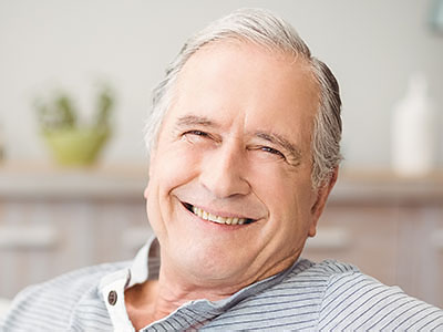The image is a photograph of an older man with gray hair, smiling gently and looking directly at the camera. He appears to be indoors, possibly in a home setting, as suggested by the domestic objects in the background.
