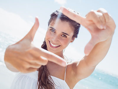 The image shows a smiling woman with long hair, wearing a white top, taking a selfie with her hands forming a frame around the camera.